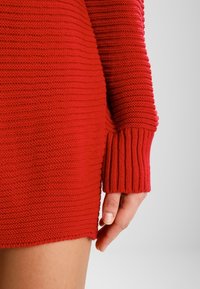 Rotes gestricktes Kleid mit gerippten Ärmeln, das ein strukturiertes Muster und einen seitlichen Schlitz aufweist. Der Stoff ist weich und hat ein engmaschiges Design.