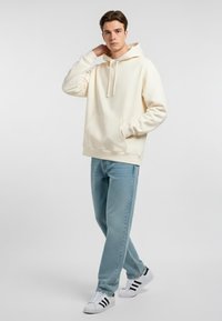 Felpa crema con tasca frontale e cappuccio con cordino. Abbinata a jeans azzurri e sneakers bianche con strisce nere.