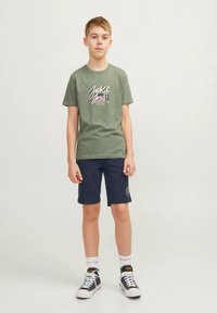 Tricou verde din bumbac cu un logo grafic, asortat cu pantaloni scurți bleumarin și șosete albe. Ținuta include adidași negri cu accente albe.