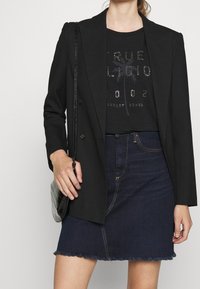 Black blazer with two buttons over a black T-shirt featuring metallic print, paired with a dark denim skirt. Textured black handbag.
