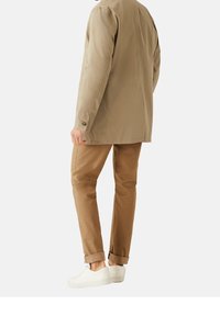 Beige leichte Jacke mit geradem Schnitt, mit Knopfmanschetten, kombiniert mit hochgekrempelten braunen Hosen und weißen Sneakers. Sauberes, minimalistisches Design.