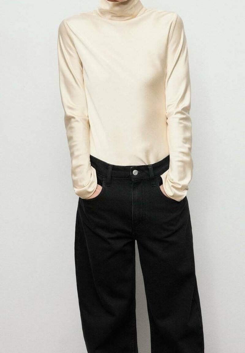 Personne portant un pull à col roulé crème à manches longues et un pantalon noir, debout les mains dans les poches devant un fond clair et uni.