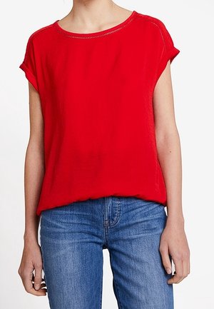 Persoon die een effen rode korte mouwen blouse draagt met opgerolde manchetten en blauwe spijkerbroek tegen een witte achtergrond.