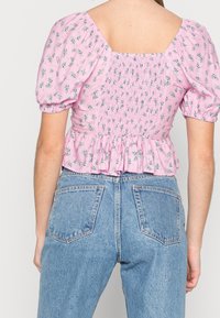 Bavlněná blůza světle růžové barvy s květinovým vzorem, krátkými volánovými rukávy, přiléhavým ráchlovým zadním dílem a nařaseným lemem, kombinovaná s modrými džínami s vysokým pasem.