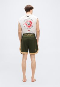 Ujjatlan fehér póló grafikával a háton, olívazöld rövidnadrággal párosítva, arany díszítéssel és márkajelzéssel. Rövid hosszúságú és könnyű anyagból készült.