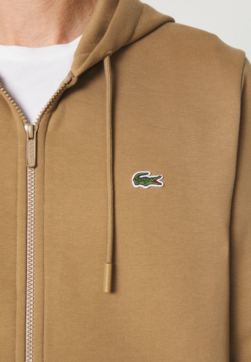 Haut à capuche zippé marron en tissu texturé, doté d'une poche kangourou à l'avant, de cordons de serrage et d'un logo de crocodile brodé en vert.