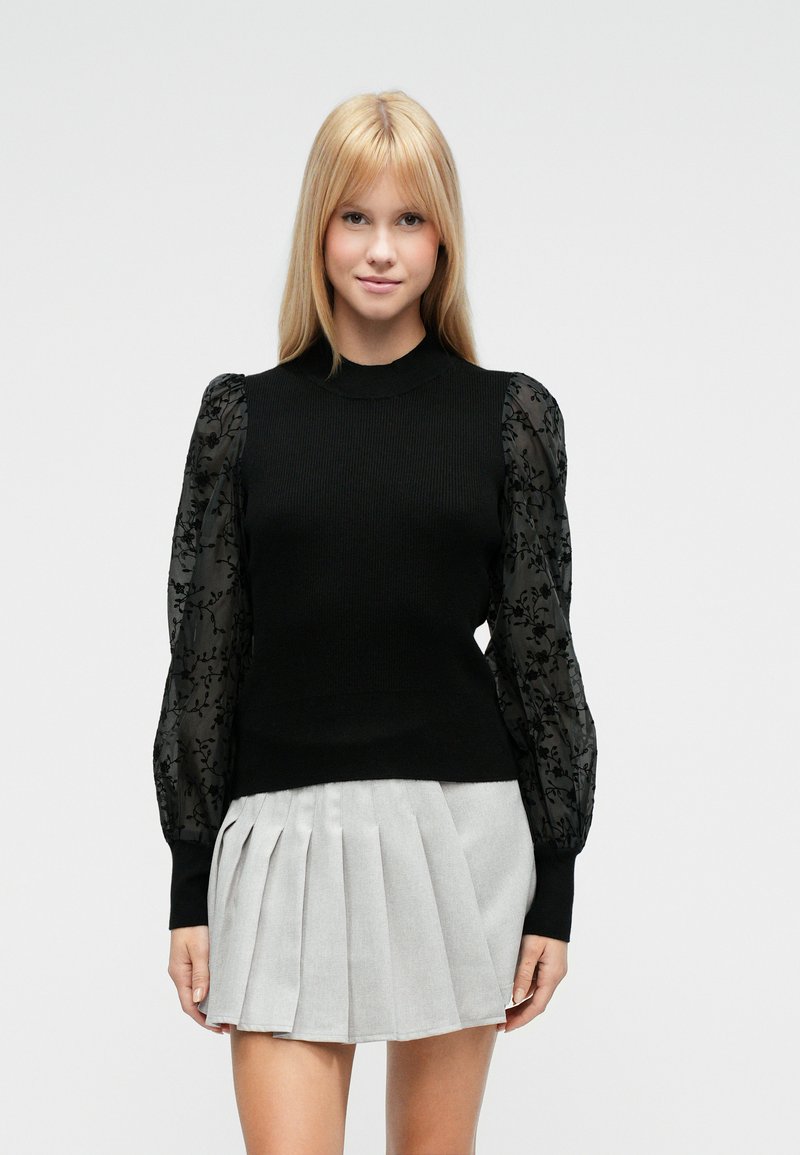 Schwarzer, gerippter Pullover mit transparenten, schwarzen Puffärmeln mit Blumenmuster, kombiniert mit einem grauen Faltenrock, der einen Kontrast aus geschichteten Texturen schafft.