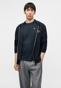 Ανδρικό ντύσιμο με μπλε σκούρο τζάκετ με φερμουάρ από υφασμάτινη υφή και κουκούλα, σε συνδυασμό με μπλε σκούρο μπλουζάκι με στρογγυλή λαιμόκοψη και ανοιχτό γκρι παντελόνι.