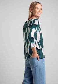 Top multicolore con un motivo astratto verde e bianco, maniche a tre quarti e una vestibilità ampia. Abbinato a jeans blu chiaro.