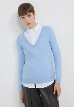 Pull en tricot bleu clair avec un col en V et des manches longues, superposé sur une chemise blanche à col. Texture douce et design ajusté.