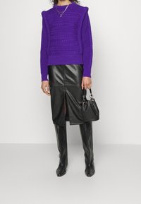 Fioletowy sweter w splot kablowy z poduszkami na ramionach, w połączeniu z czarną skórzaną spódnicą midi z rozcięciem z przodu i wysokimi czarnymi botkami.