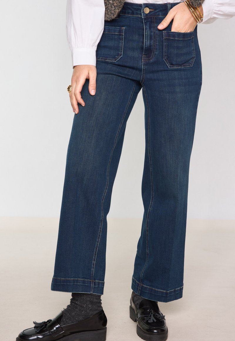 Kvinde iført mørkeblå jeans med vide ben og frontlommer, sorte loafers, mørke sokker, hvid bluse og gyldne armbånd.