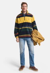 Mann trägt einen mehrfarbigen gestreiften Fleece-Pullover, helle blaue Jeans, braune Schuhe und hält eine gefaltete beige Jacke vor einem weißen Hintergrund.