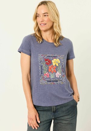Woman with blonde hair wearing a blue t-shirt with colorful embroidered flowers and text, paired with blue jeans, against a plain background.