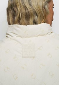 Gepolsterte cremefarbene Jacke mit Rautenmuster und hellbeigen "10" Logos, hohem Kragen und rechteckigem Logoaufnäher auf der Rückseite.