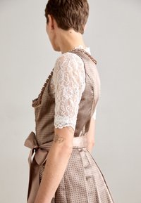 Femme aux cheveux courts portant une robe beige à motifs avec un nœud en satin et des manches en dentelle florale blanche, montrant des tatouages sur son bras.