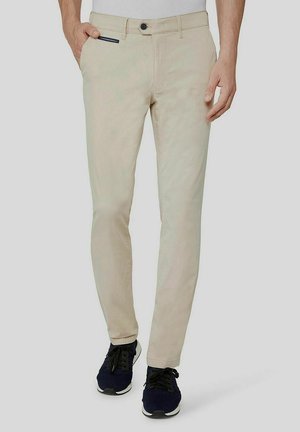 Man draagt beige slim-fit broek met zwarte sneakers, één hand in de zak tegen een effen lichtgrijze achtergrond.