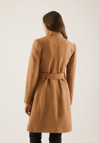 Kobieta z długimi brązowymi włosami, ubrana w wełniany płaszcz koloru wielbłądziego, z paskiem, widziana od tyłu na jednolitym tle.