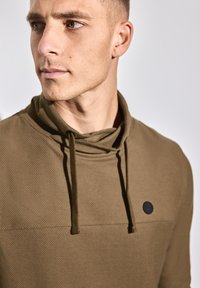 Olivově zelená mikina s texturovaným povrchem, vybavená kapucí a šňůrkami, s černým kruhovým logem na hrudi.