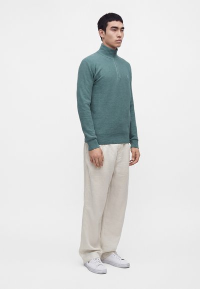 Homme portant un pull zippé bleu sarcelle, un pantalon blanc ample et des baskets blanches, debout devant un fond clair uni.