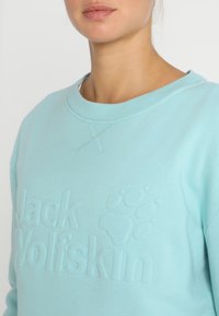 Světle modrá mikina s vyraženým logem "Jack Wolfskin" a otiskem tlapky. Klasický design s kulatým výstřihem a jemnými švy.