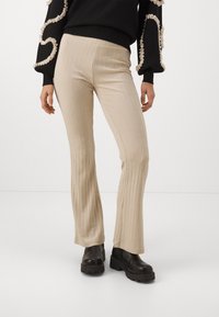 Pantalones acampanados de canalé beige con un acabado texturizado, que cuentan con una cintura ajustada y una cómoda elasticidad. Llevados con botas negras de suela gruesa.