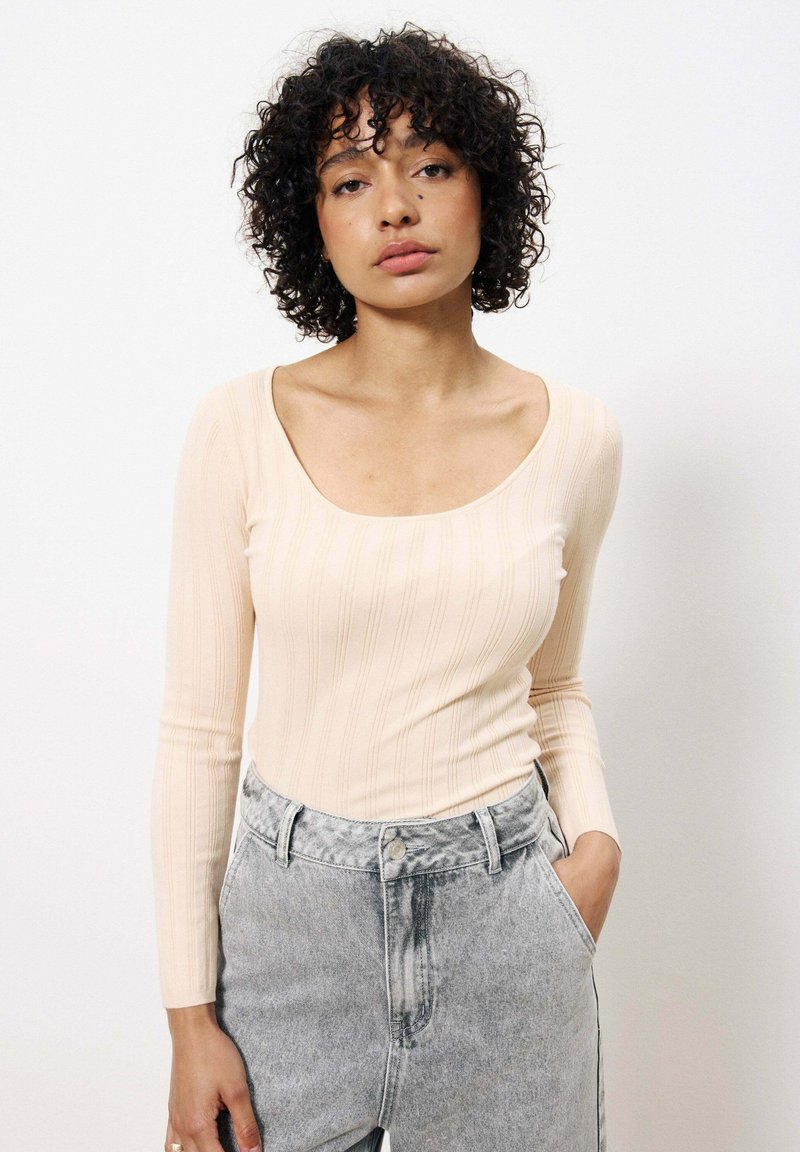 Top long à manches longues beige clair côtelé avec un col rond large, associé à un jean gris taille haute. Design simple et texturé.