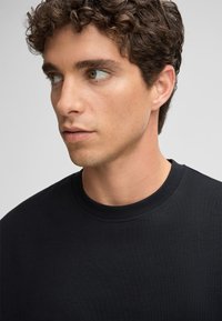 Schwarzes Baumwoll-T-Shirt mit rundem Halsausschnitt und kurzen Ärmeln, an einem Modell präsentiert. Glatte Textur und standardmäßige Passform sichtbar.
