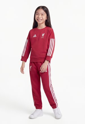 Junges Mädchen lächelt, trägt einen roten Liverpool FC Trainingsanzug mit weißen Adidas-Streifen und weißen Turnschuhen, steht vor einem weißen Hintergrund.