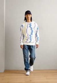 Dzianinowy sweter o białej bazie, z niebieskimi, fioletowymi i żółtymi wzorami w zygzak. Noszony z niebieskimi dżinsami i białymi sneakersami.