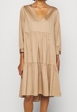 Vrouw draagt een loszittende, beige midi-jurk met lange mouwen en V-hals, voorzien van geplooide lagen en met knoopmanchetten, weergegeven van nek tot knieën.