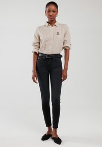 Žena u bež prugastoj košulji s gumbima, uvučenoj u crne uske farmerke s visokim strukom, crnim remenom i crnim slip-on cipelama.