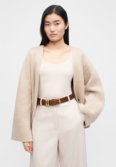 Cardigan bej tricotat, cu mâneci largi, peste un top ușor. Asortat cu pantaloni crem cu talie înaltă și o curea maro cu cataramă aurie.