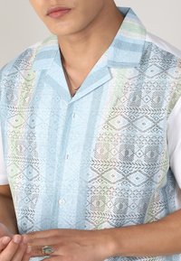 Chemise à manches courtes de couleur bleu clair avec des motifs géométriques, dotée d'une fermeture à boutons et d'accents dégradés de couleur verte et blanche.