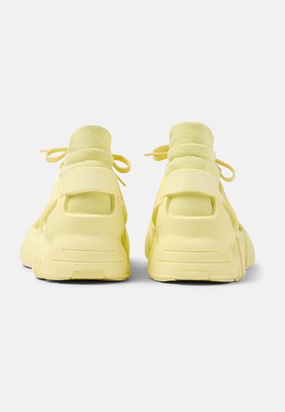 Sneaker gialli con colletto imbottito, tomaia in mesh, lacci decorativi e suola in gomma testurizzata, che presenta un design arrotondato e robusto sul retro.