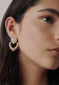 Gold-colored heart-shaped earrings with a textured twisted design, displayed on a model's ear. Smooth skin tone in the background.