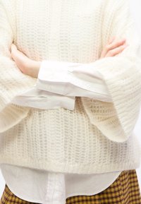 Άτομο με σταυρωμένα χέρια που φοράει λευκό πουλόβερ με υφή πάνω από λευκό πουκάμισο και καφέ παντελόνι καρό.