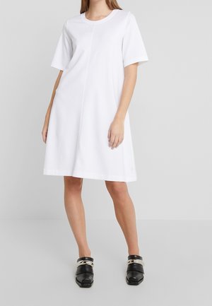 Kobieta w białej sukience do kolan z krótkimi rękawami i czarnych wsuwanych butach z metalicznymi akcentami, stojąca na jednolitym szarym tle.