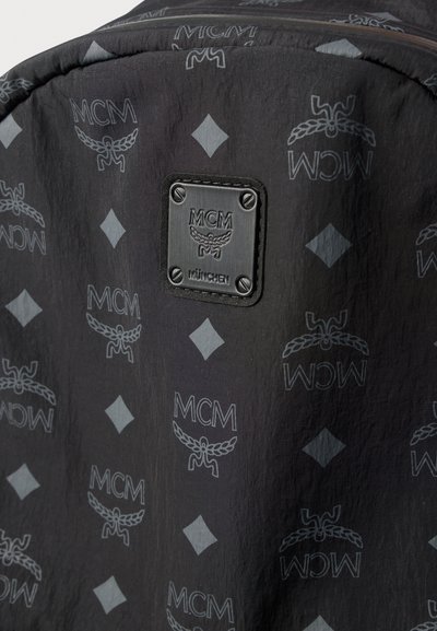 Mochila de tecido preto com um padrão tonal subtil com logos MCM e formas de diamante. Destaca-se uma placa de metal preta com o logo.