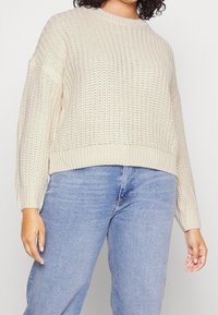Kobieta w kremowym, dzianym swetrze z bufiastymi rękawami oraz jasnoniebieskich, wysokogatunkowych dżinsach, stojąca na jednolitym tle.