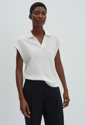 Vrouw die een mouwloze witte gebreide top met kraag en zwarte hoge taillebroek draagt, staand tegen een eenvoudige lichte achtergrond.