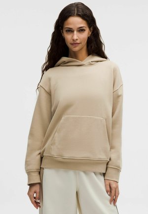 Kobieta o długich ciemnych włosach, ubrana w beżową bluzę z kapturem i kieszenią z przodu oraz jasne spodnie, stoi na tle jednolitego tła.