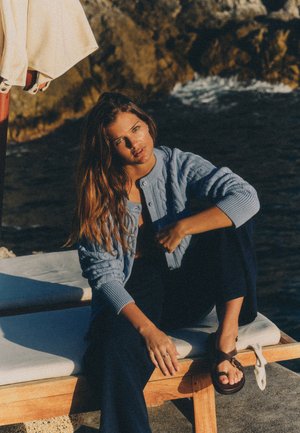 Noor naine, kellel on pikad juuksed, sinise kootud kardigani ja tumedate pükstega, istub puidust pingil kivisel kaldal päikeseloojangul.