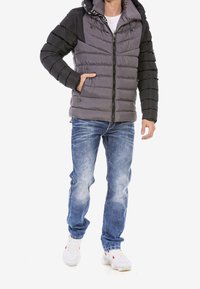 Chaqueta acolchada gris y negra con cremallera, capucha y cordones, combinada con jeans azules desgastados y zapatillas blancas.