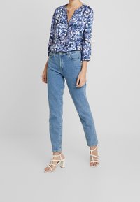 Niebieska bluzka w wzory z długimi rękawami, zestawiona z jasnoniebieskimi spodniami o podwyższonym stanie oraz białymi sandałami na słupkowym obcasie z paskami.