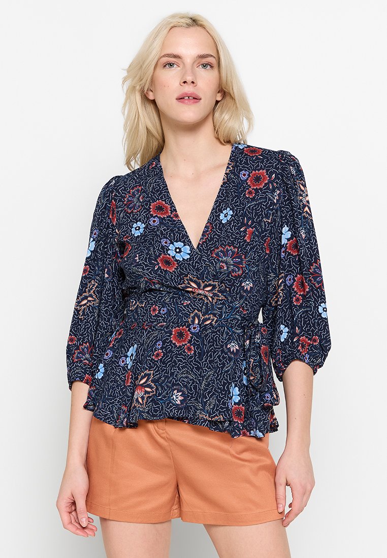 Blusa floral con escote en V profundo, fondo azul marino y flores rojas y azules. Presenta mangas largas abullonadas y un dobladillo tipo peplum.
