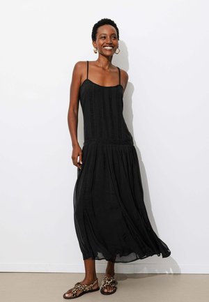 Femme souriante portant une robe midi noire sans manches et des sandales à imprimé léopard, debout contre un mur blanc.