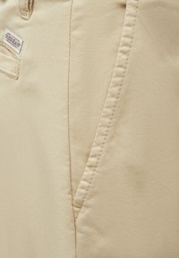 Gros plan sur un pantalon en tissu beige avec des coutures visibles, une poche avant, un passants de ceinture et une petite étiquette rectangulaire Guess Jeans près de la poche.