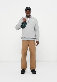 Felpa grigia con zip, pantaloni beige e scarpe nere. Gli accessori includono un cappellino nero e una borsa a tracolla verde. Design semplice e casual.