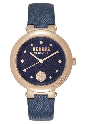 Rund guldtonet Versus Versace-ur med blå tekstureret urskive, gyldne markører, løveemblem ved klokken 6, og blå tekstureret læderrem.